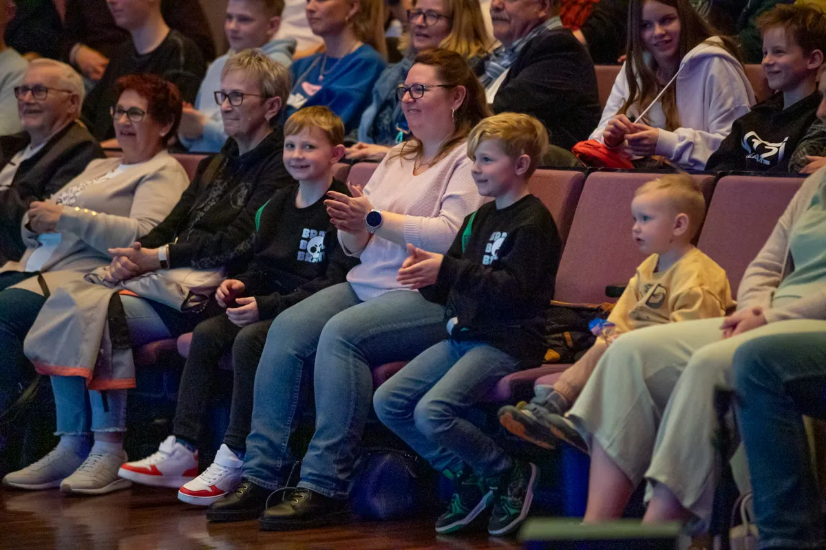 Zuschauer sitzen auf roten Sitzen in einem Theater und beobachten eine Vorstellung. Im Vordergrund ein lächelndes Kind, das in die Hände klatscht, neben einer Frau mit Brille.