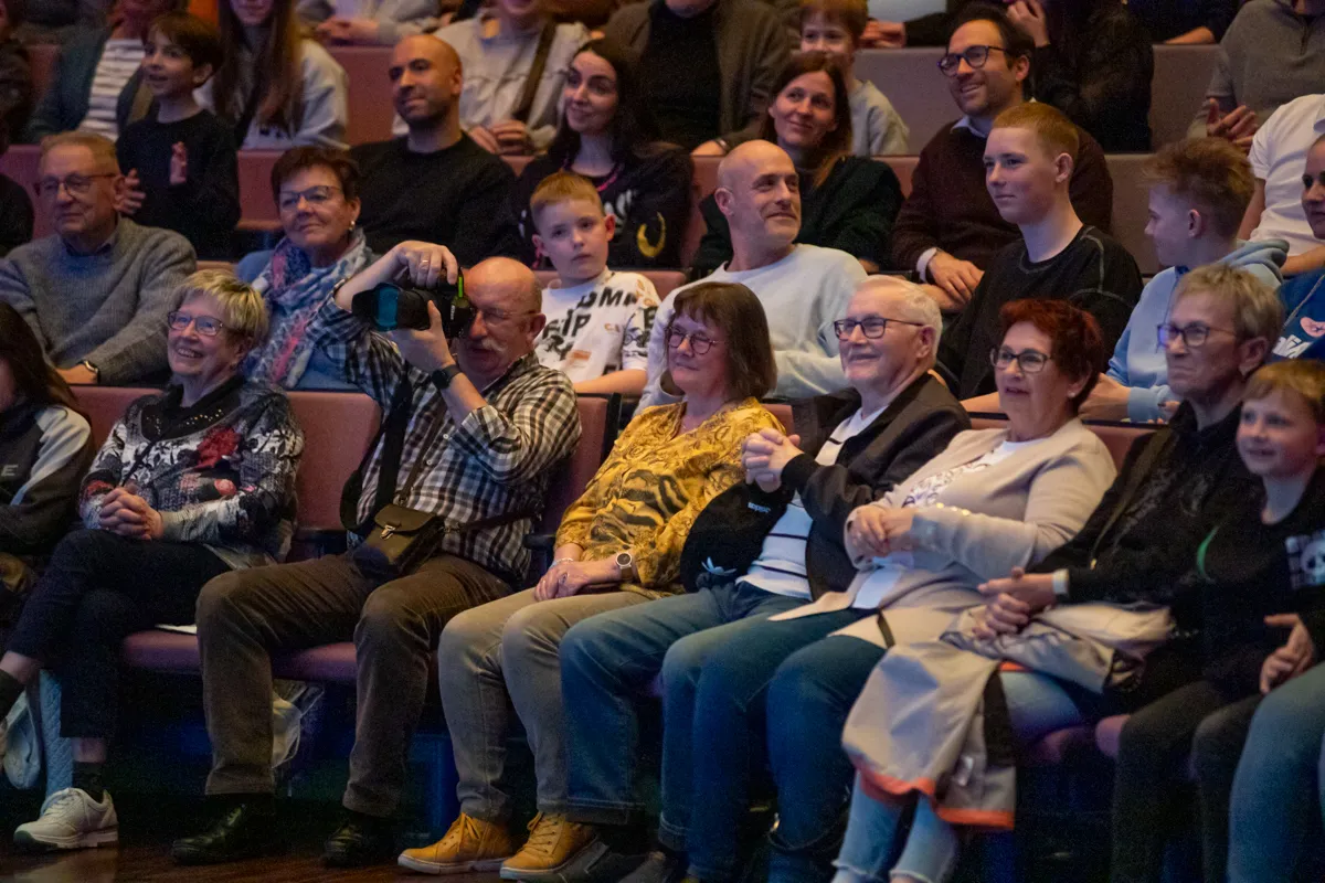 Zuschauer sitzen auf roten Sitzen in einem dunklen Theatersaal und schauen auf die Bühne. Im Vordergrund eine Frau mit grauen Haaren und Brille, neben ihr eine ältere Dame mit rotem Pullover.