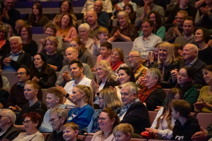 Zuschauer klatschen und lächeln während einer Vorstellung im Theater. Im Vordergrund eine Frau mit blauem Pullover und hochgestecktem Haar.
