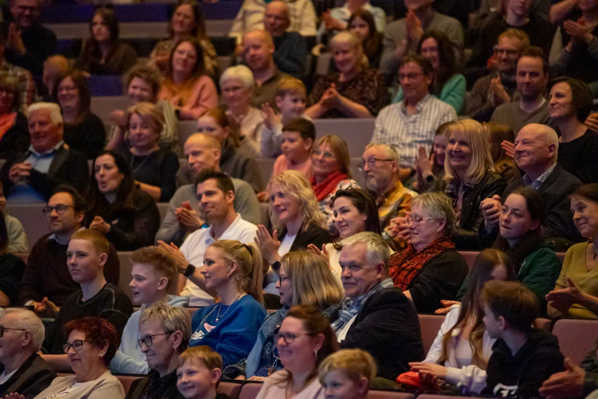 Zuschauer klatschen und lächeln während einer Vorstellung im Theater. Im Vordergrund eine Frau mit blauem Pullover und hochgestecktem Haar.