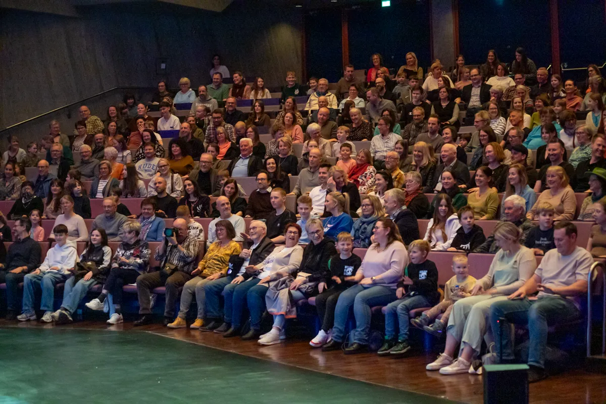 Zuschauer sitzen auf roten Sitzen in einem Theater und schauen in Richtung Bühne. Die Reihen sind voll besetzt mit Menschen unterschiedlichen Alters.