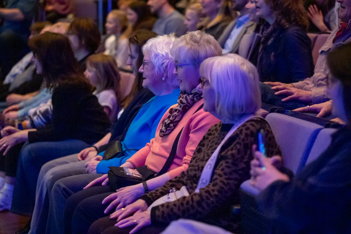 Zuschauer sitzen auf blauen Sitzen in einem dunklen Raum und schauen auf eine Bühne. Mehrere ältere Frauen sind im Vordergrund zu sehen.
