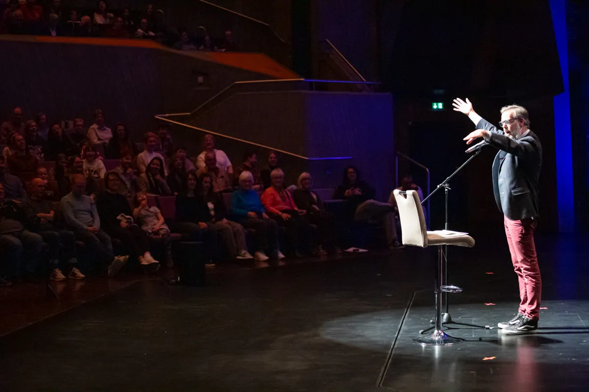 Ein Mann mit Brille und im Blazer gestikuliert auf einer Bühne vor einem Publikum in einem Theater. Neben ihm steht ein Stuhl vor einem Mikrofonständer.
