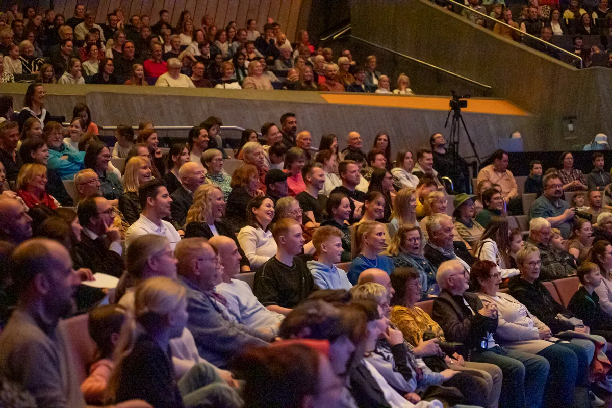 Zuschauer sitzen in einem dunklen Saal auf roten Sitzen und schauen auf eine Bühne. Viele Menschen unterschiedlichen Alters und Geschlechts sind zu sehen.