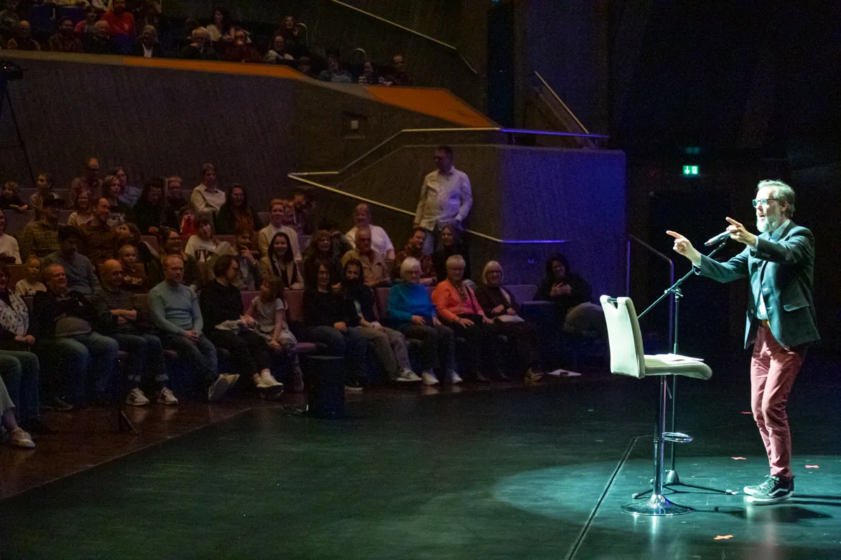 Ein Mann mit Bart und Brille gestikuliert auf einer Bühne vor einem Publikum. Im Hintergrund sind die Ränge eines Theaters mit Zuschauern gefüllt.