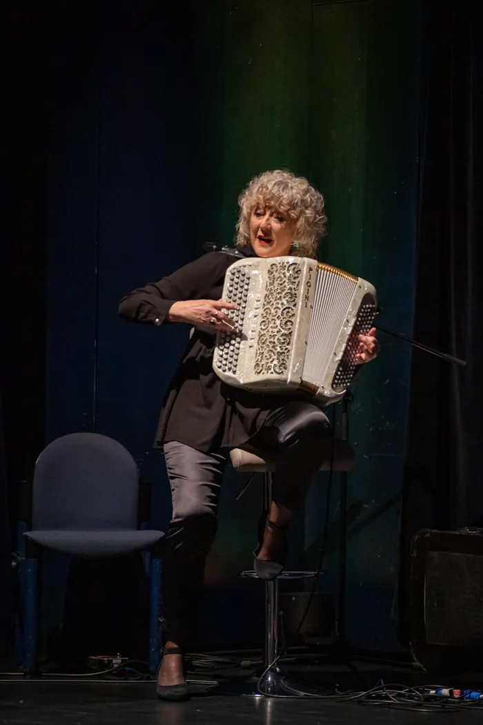 Eine Frau mit gelocktem, grauem Haar spielt auf einem weißen Akkordeon, während sie auf einem Barhocker sitzt. Sie trägt ein schwarzes Outfit und scheint auf einer Bühne zu stehen.