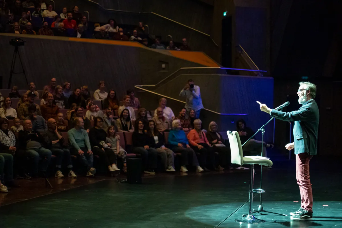 Ein Mann mit Bart hält ein Mikrofon auf einer Bühne vor einem Publikum in einem Theater. Die Zuschauer sitzen auf Stühlen und schauen ihm zu.