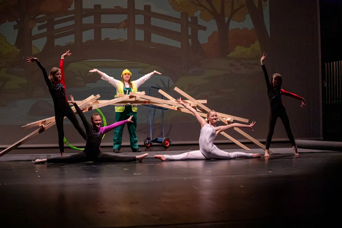 Fünf junge Tänzer in verschiedenen Posen auf einer dunklen Bühne, einige halten Holzlatten. Im Hintergrund eine Kulisse mit Bäumen und einem Zaun.