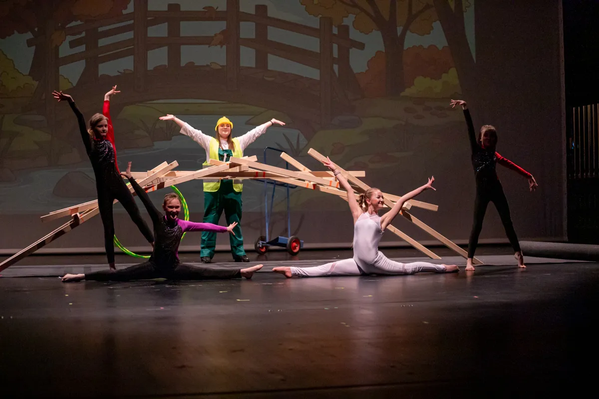 Fünf Tänzer in Bühnenkostümen und ein Kind mit Bauarbeiterhelm und Weste posieren vor einem Bühnenbild mit Baum- und Zaunmotiven. Einige Tänzer halten Holzstäbe.