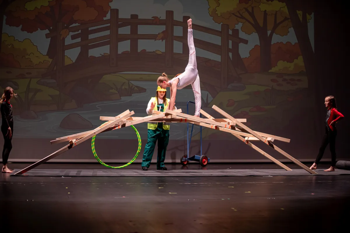 Eine Akrobatin balanciert auf gestapelten Holzstangen, während eine Person in Arbeitskleidung mit gelbem Helm darunter steht. Zwei weitere Darsteller stehen seitlich auf der Bühne vor einer Kulisse mit Landschaftsdarstellung.
