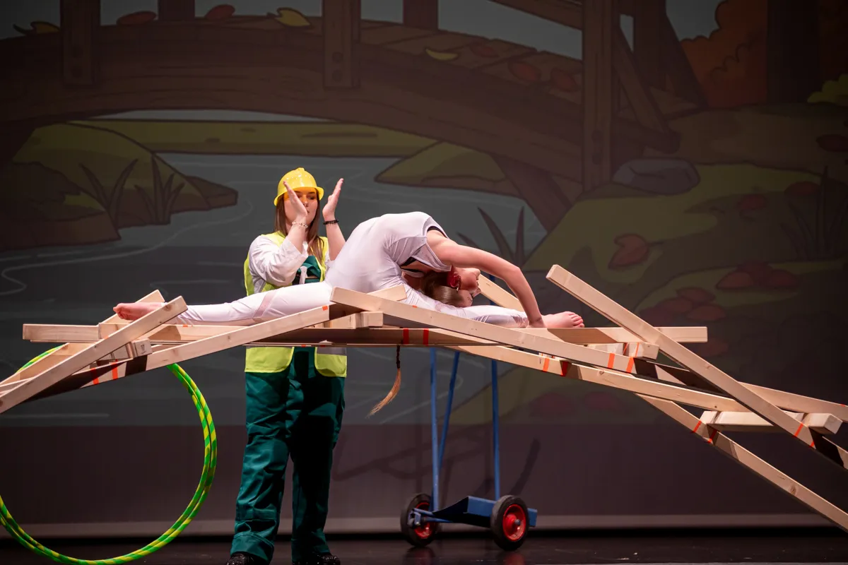 Eine Akrobatin balanciert auf einem Gerüst aus Holzlatten, während eine Person mit gelber Bauarbeiterkappe und grüner Kleidung ihre Hand ausstreckt. Im Hintergrund eine Kulisse mit Waldmotiven.