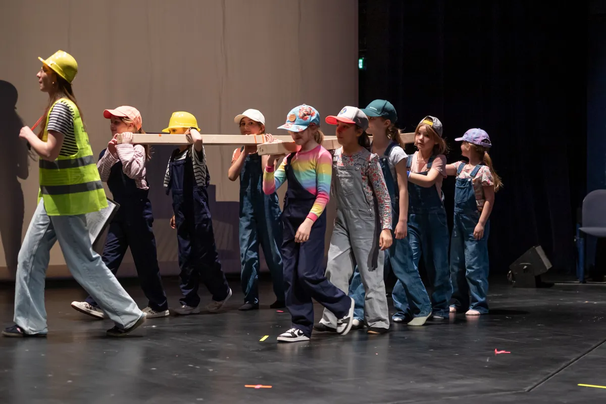 Eine Gruppe von Kindern und Jugendlichen in Arbeitskleidung und Helmen trägt auf einer Bühne ein langes Holzbrett. Eine Person mit Warnweste führt die Gruppe an.