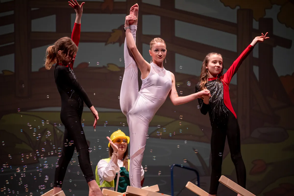 Vier junge Artisten in Bühnenkostümen performen eine akrobatische Übung vor Publikum. Eine trägt einen gelben Hut und macht ein Foto.