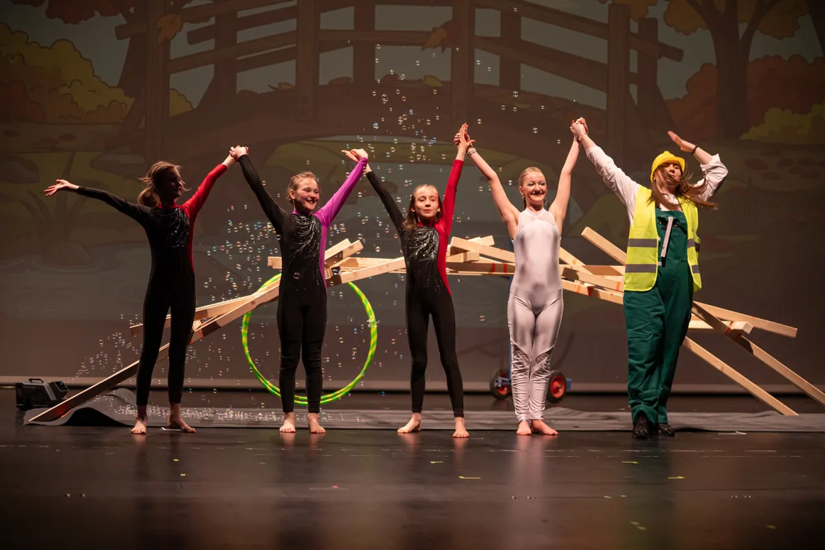 Fünf Kinder stehen auf einer Bühne mit erhobenen Armen, einige tragen Clowns-Kostüme, eines einen Overall mit Helm. Im Hintergrund eine Kulisse mit Bäumen und schwebende Seifenblasen.