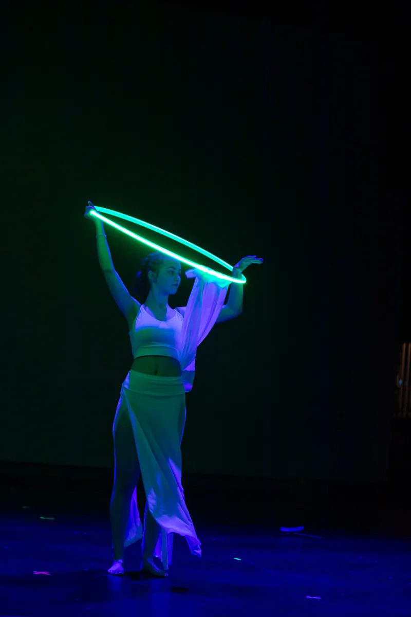 Tänzerin in weißem Kostüm hält einen leuchtenden Reifen über den Kopf, während sie auf einer dunklen Bühne steht.