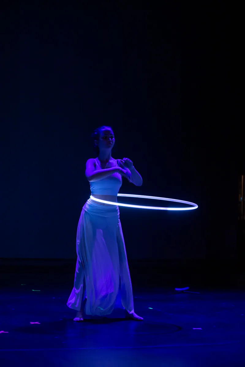 Eine Frau in weißem Kleid und Oberteil dreht auf einer Bühne einen leuchtenden Reifen. Blaue Beleuchtung erzeugt eine dramatische Atmosphäre.