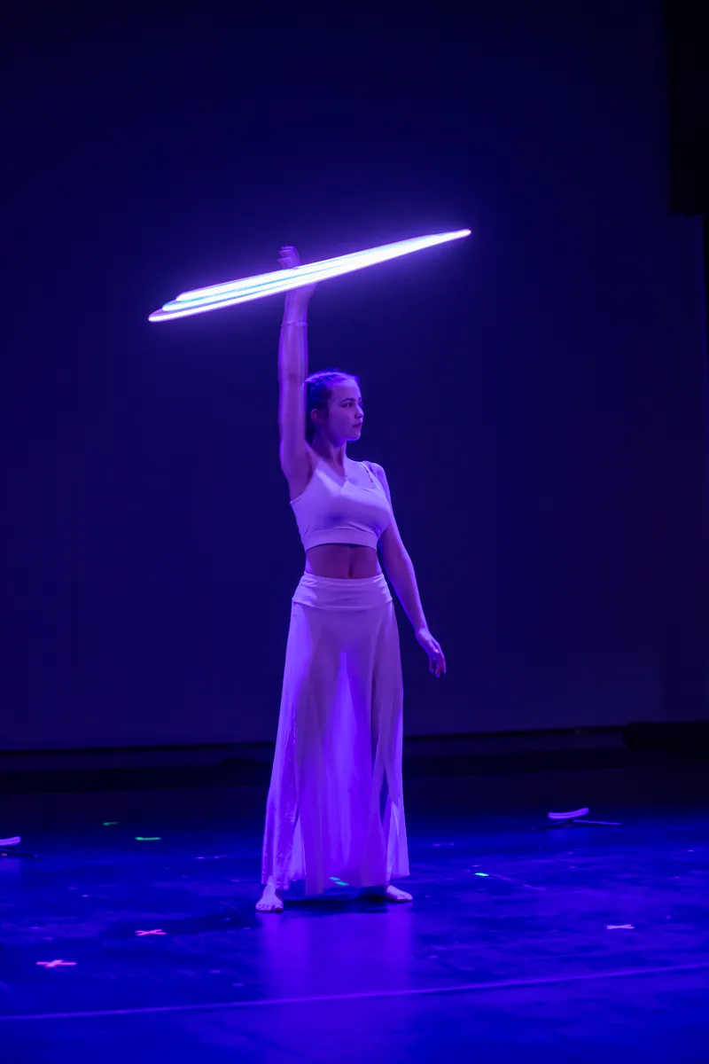 Tänzerin in weißem Outfit hält leuchtendes Schwert über dem Kopf, während sie auf einer dunklen Bühne steht.