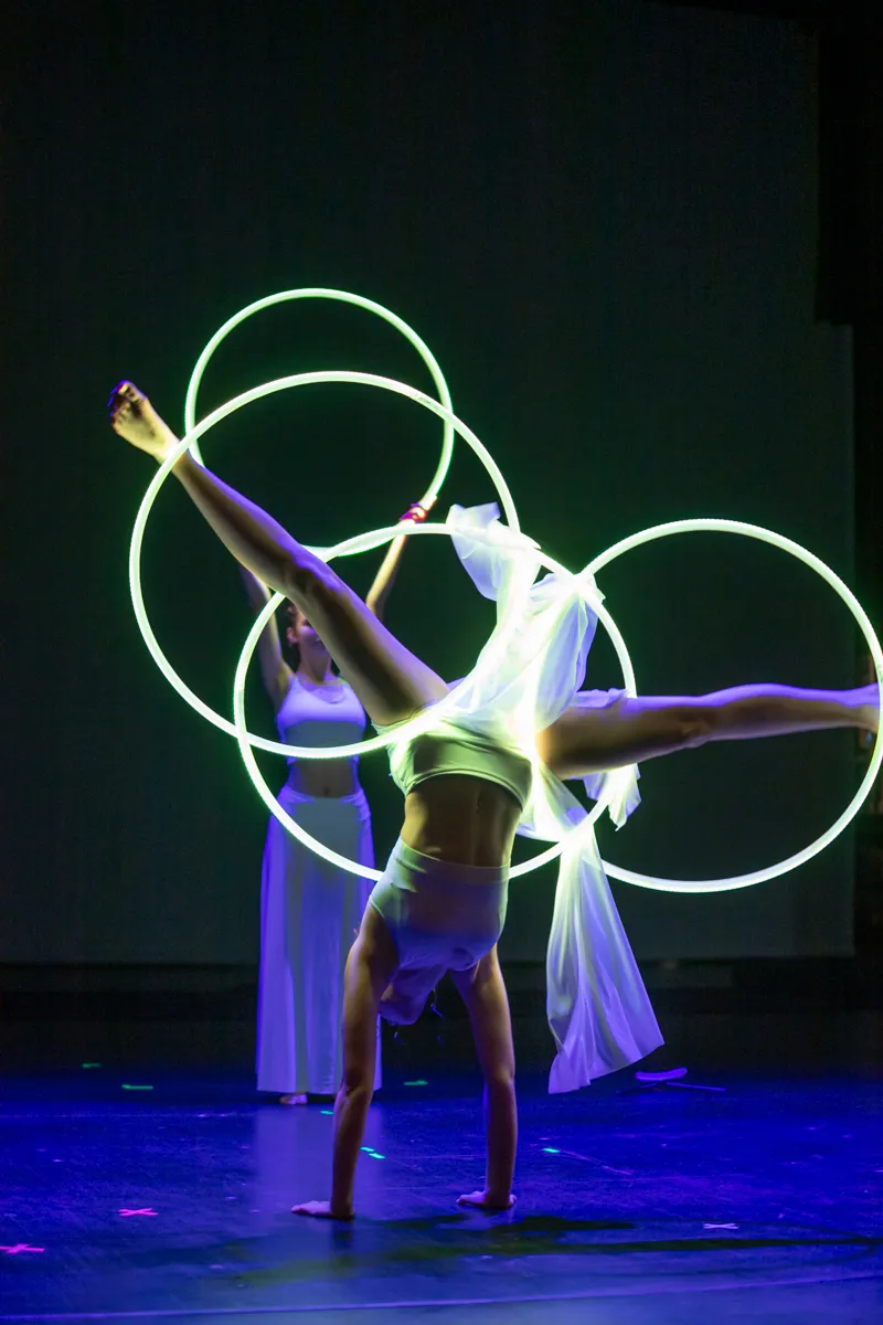 Eine Akrobatin hält in einem Handstand mit leuchtenden Reifen, während eine zweite Person im Hintergrund steht.