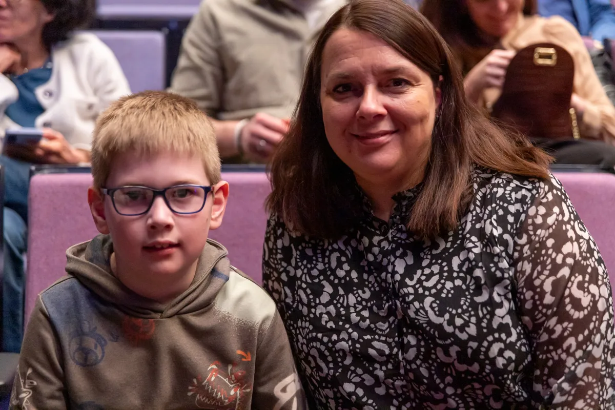 Junge mit Brille und Frau lächeln in einem Theater. Im Hintergrund sind weitere Zuschauer zu sehen.