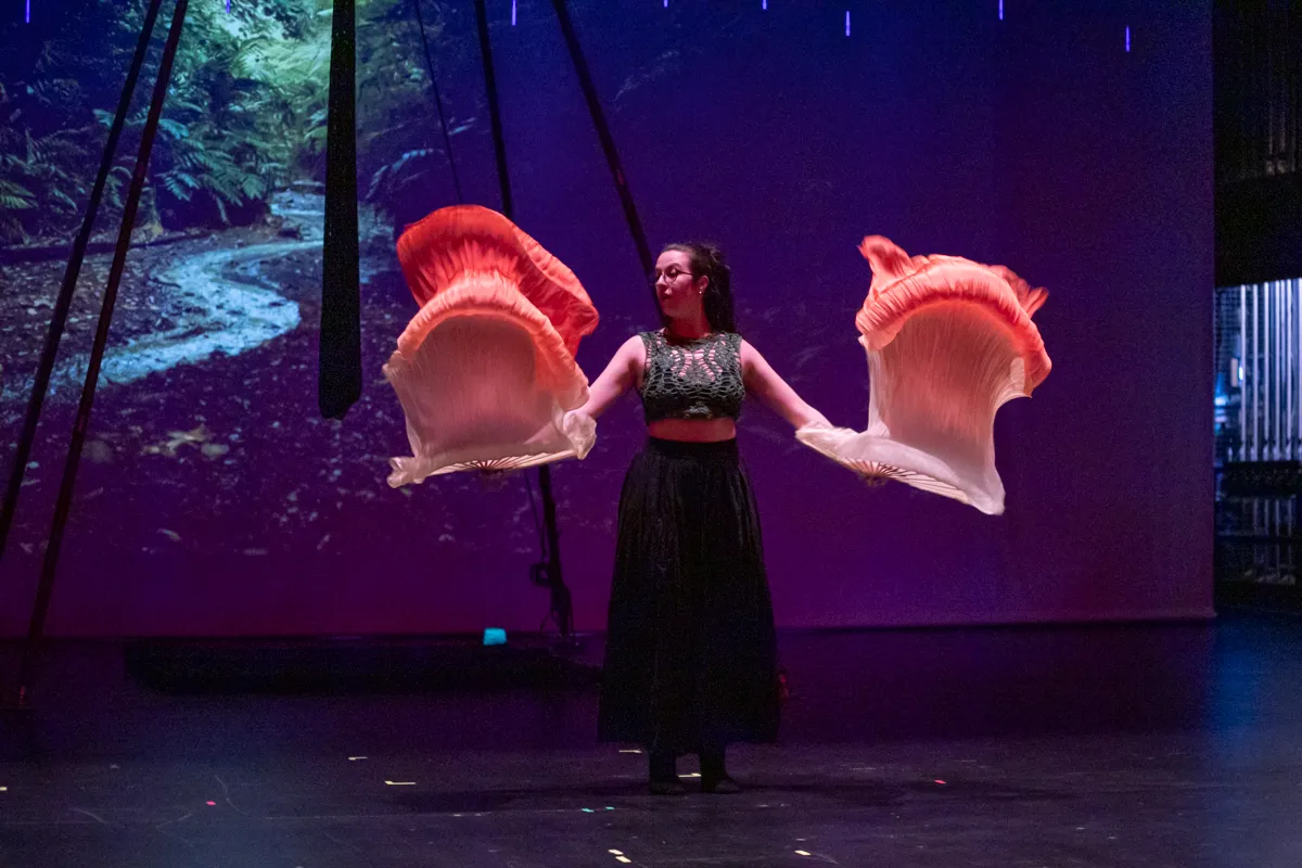 Eine Frau mit langen braunen Haaren und Brille tanzt auf einer Bühne mit großen, orange-weißen Fächern in den Händen. Im Hintergrund eine dunkle Waldkulisse und violettes Licht.