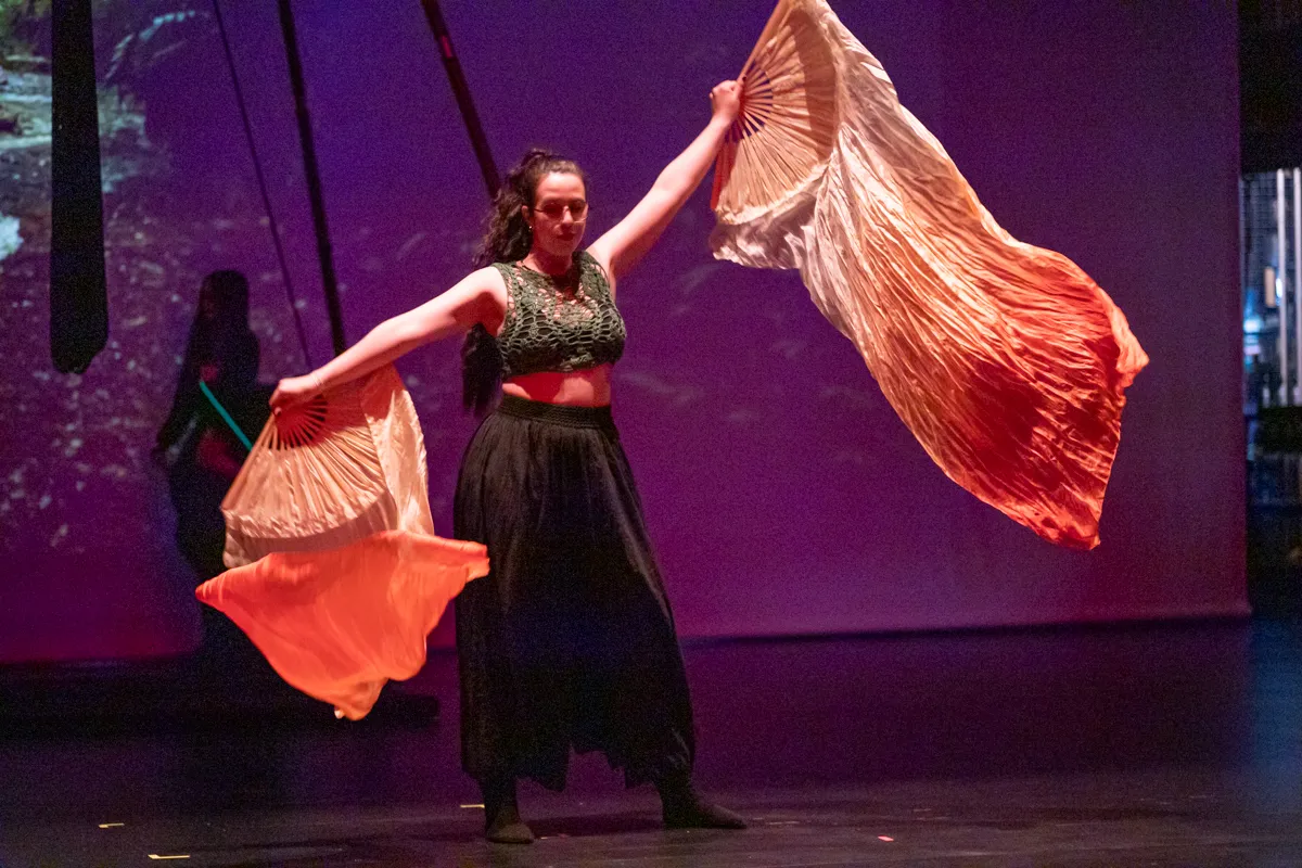 Eine Tänzerin in einem grünen Oberteil und einem langen schwarzen Rock wirbelt mit orangefarbenen Fächern auf einer violett beleuchteten Bühne. Im Hintergrund ist eine weitere Person zu sehen.