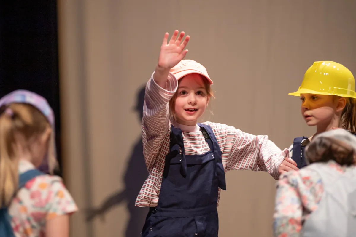 Ein Mädchen mit rosa Kappe und blauem Overall winkt auf einer Bühne. Im Hintergrund sind weitere Kinder in Kostümen zu sehen, eines trägt einen gelben Helm.
