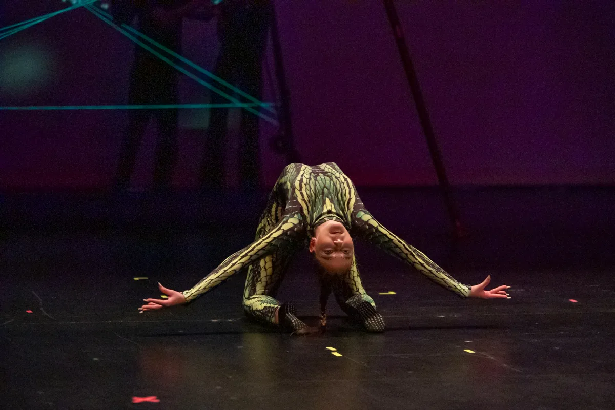 Eine Tänzerin in einem grünen und gelben Ganzkörperkostüm beugt sich auf der Bühne nach vorne, die Arme sind seitlich ausgestreckt. Im Hintergrund sind blaue Lichtinstallationen zu sehen.