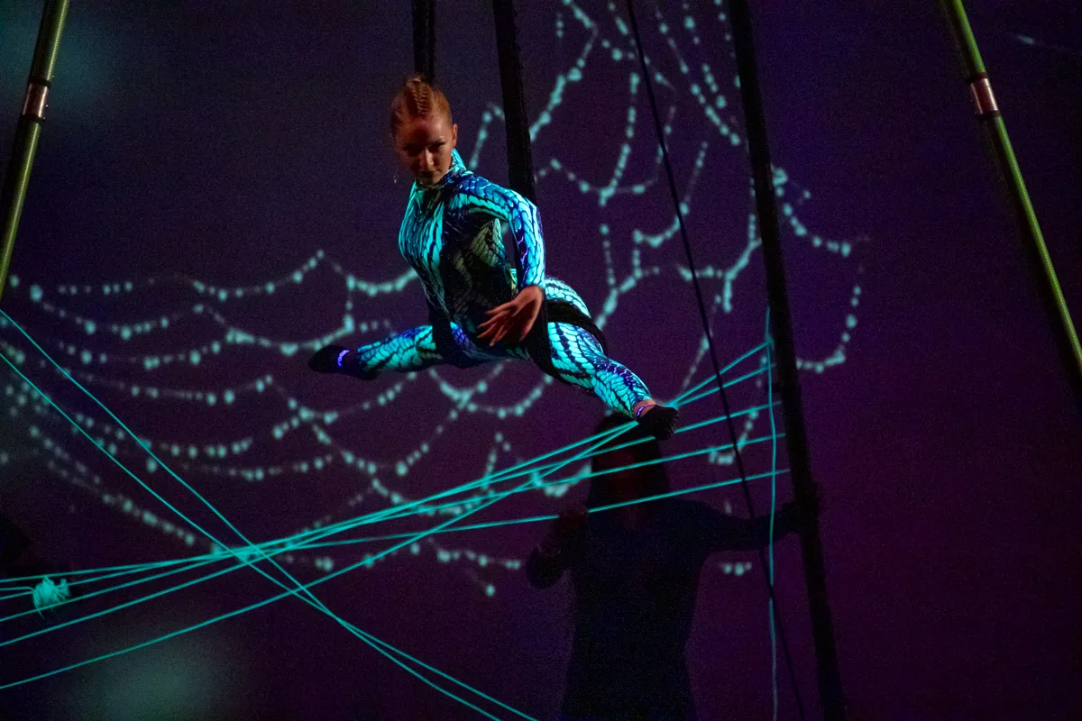 Eine Akrobatin in einem blauen Ganzkörperkostüm schwebt zwischen Seilen in der Luft, umgeben von leuchtenden, blauen Lichtlinien.