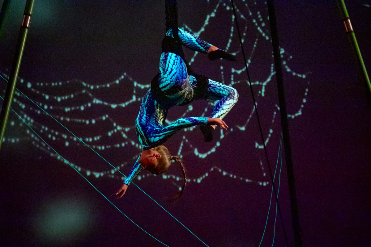 Eine Akrobatin in blauem und türkisfarbenem Kostüm balanciert an einem Seil, das zwischen zwei Stangen gespannt ist. Im Hintergrund ist ein dunkler Raum mit hellen Linien zu sehen.