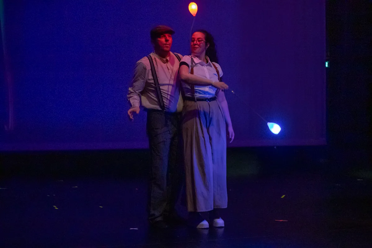 Zwei Darsteller in Kostümen mit Hosenträgern und Hüten stehen auf einer Bühne vor blauem Hintergrund und halten leuchtende Ballons.