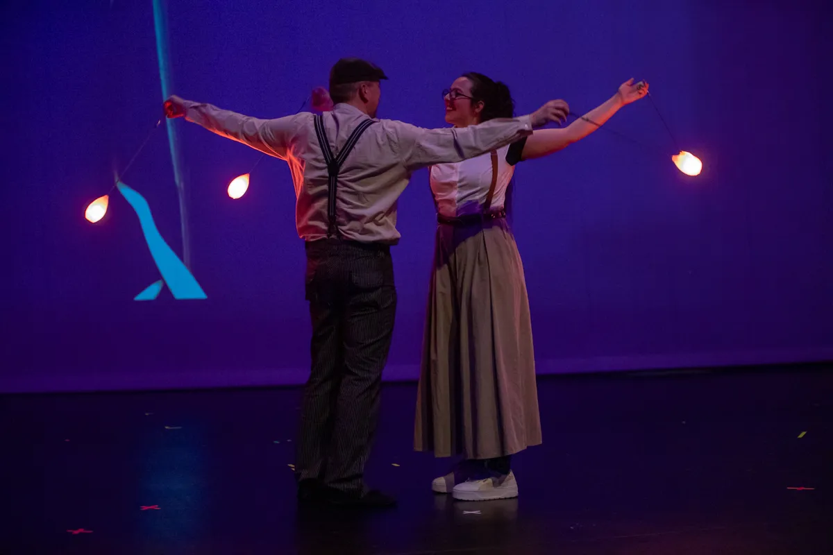 Zwei Artisten mit Hosenträgern jonglieren auf einer Bühne mit roten Bällen, während sie sich im Tanz zuwenden.