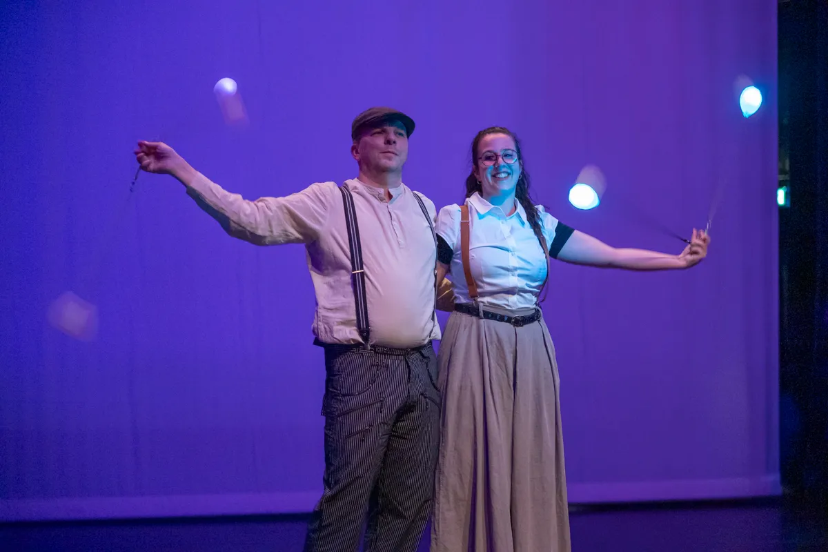 Zwei Darsteller in Kostümen mit Hosenträgern und Hüten jonglieren auf einer Bühne vor blauem Hintergrund.