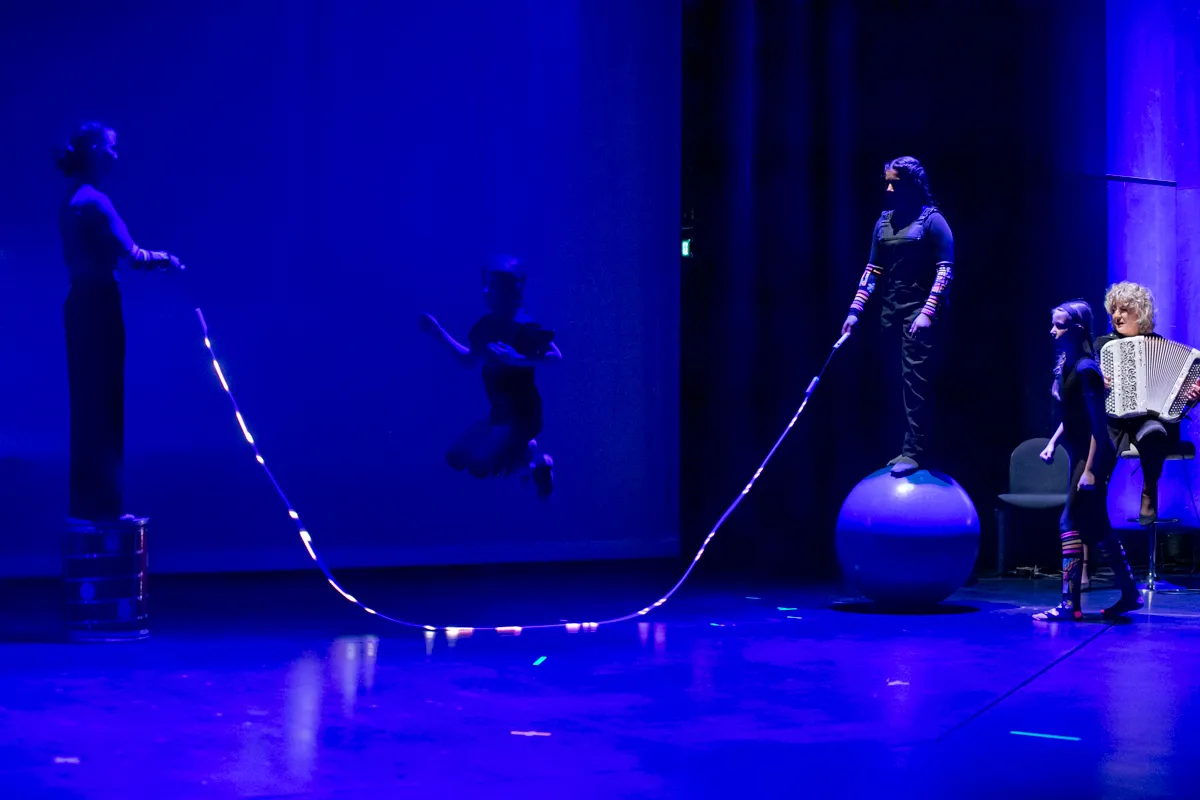 Vier Darstellende auf einer Bühne: Eine Person springt über ein Seil, eine andere spielt Akkordeon, während zwei weitere das Seil halten und balancieren.