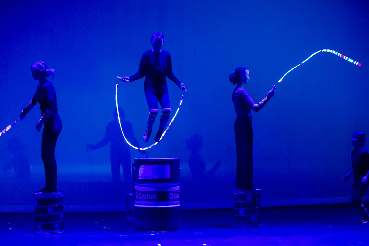 Eine Akrobatin springt über ein leuchtendes Seil, das von zwei weiteren Darstellerinnen gehalten wird, während sie auf Fässern stehen. Im Hintergrund sind weitere Personen zu sehen.