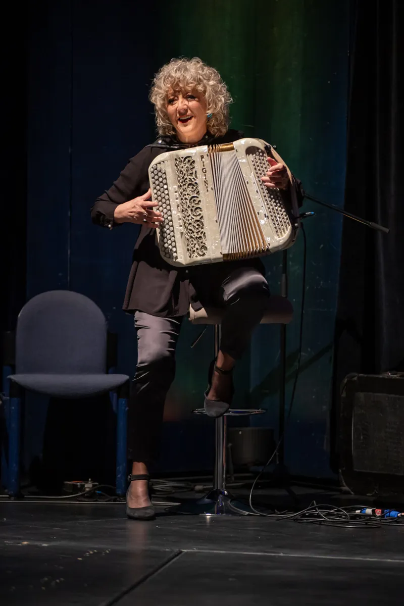 Eine Frau mit lockigem, grauem Haar spielt auf einem Akkordeon auf einer Bühne. Sie lächelt und trägt ein schwarzes Outfit.