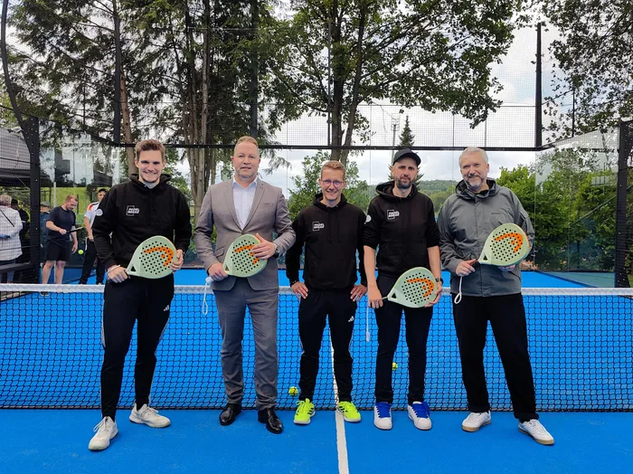 Fünf Männer stehen auf einem Padel-Tennisplatz. Drei von ihnen tragen schwarze Hoodies mit dem Logo 
