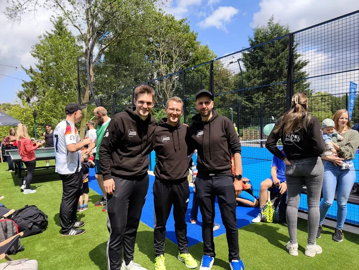 Kevin Krieter, Marcel Krings und Danijel Cvjetkovic stehen vor einem Padel-Tennisplatz. Alle drei tragen schwarze Jacken mit dem Aufdruck 