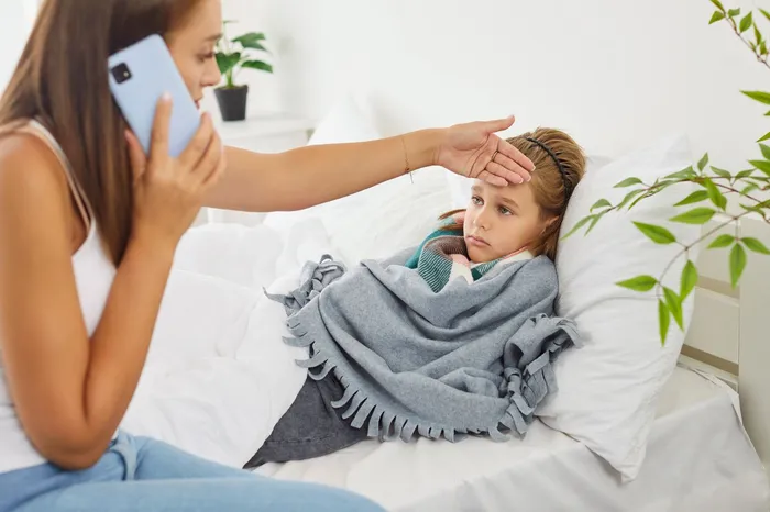 Eine Frau misst mit der Hand die Temperatur eines erkrankten Kindes, das im Bett liegt und besorgt dreinblickt. Im Hintergrund ist ein Smartphone zu sehen.