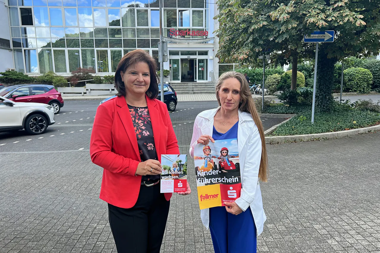 Manuela Keiderling und Susanne Fellmer präsentieren Flyer für die Kinderführerschein-Aktion vor einem Sparkasse-Gebäude.