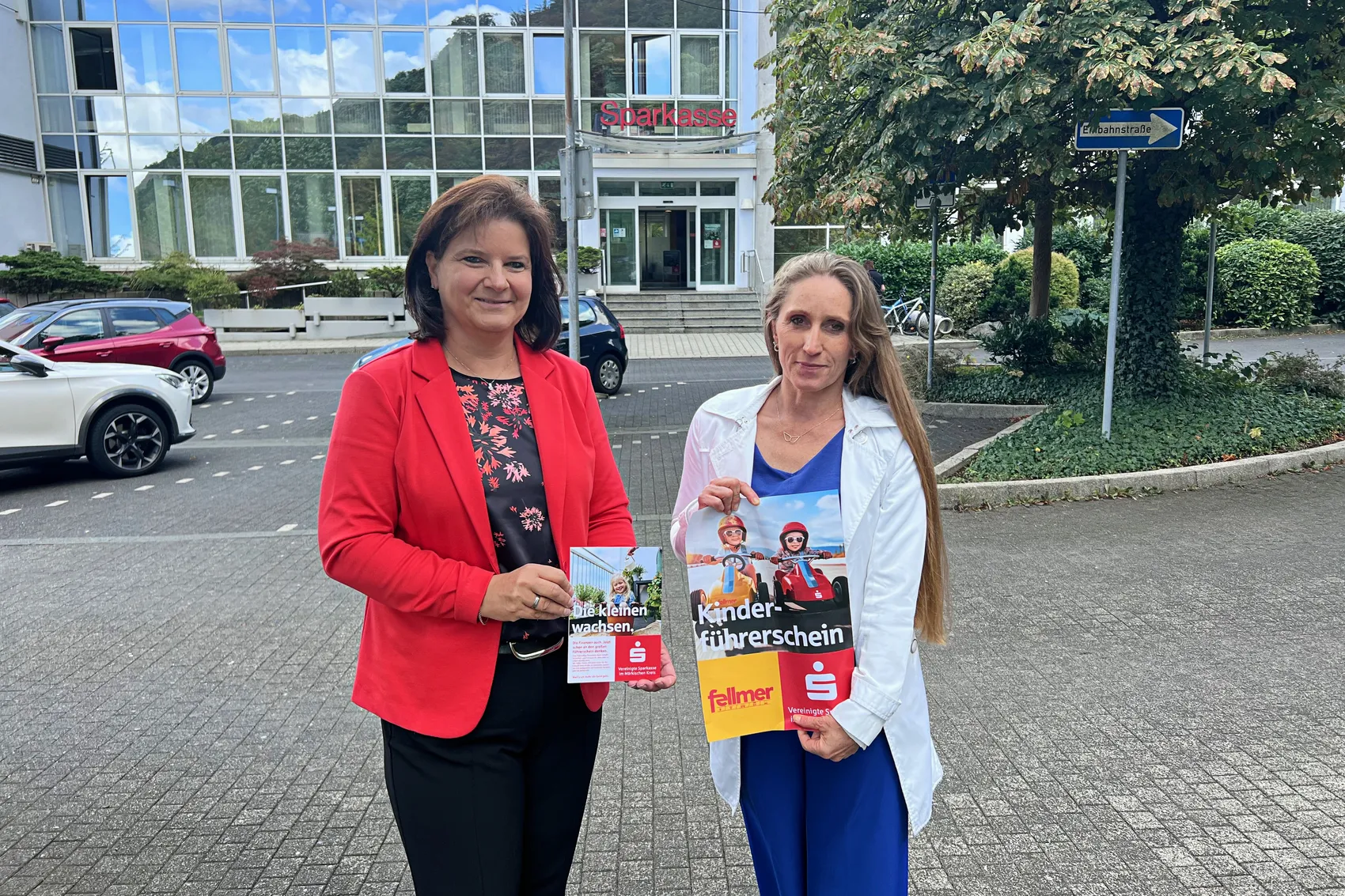 Manuela Keiderling und Susanne Fellmer präsentieren Flyer für die Kinderführerschein-Aktion vor einem Sparkasse-Gebäude.