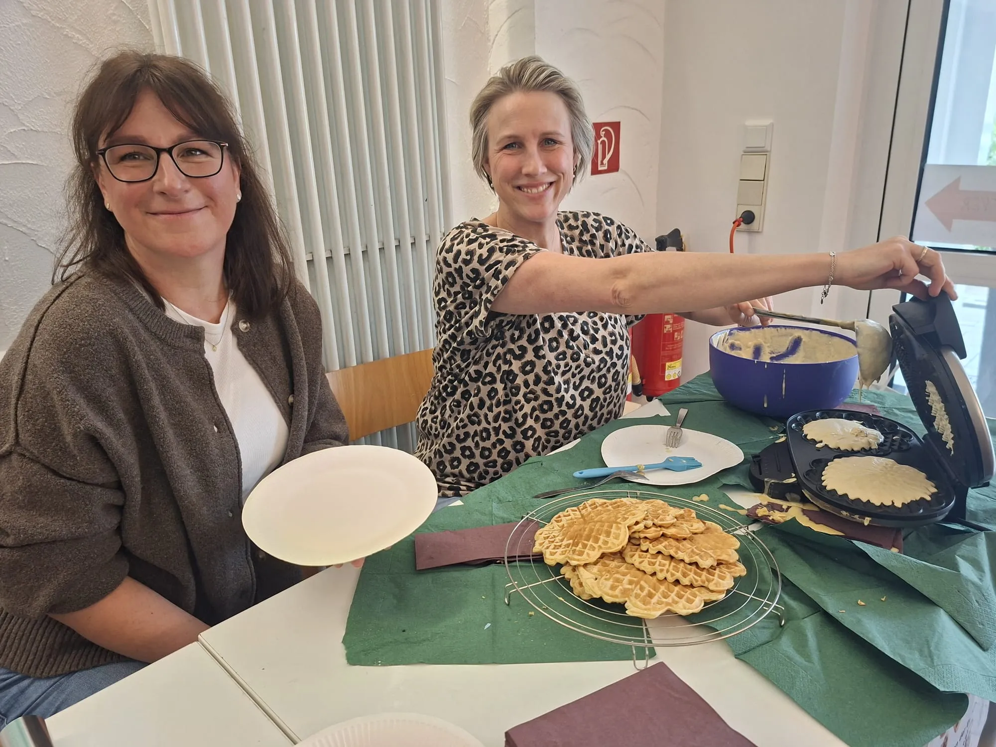 Zwei Frauen stehen an einem Tisch mit Waffeln und Zubehör. Eine Frau hält ein Waffeleisen, die andere lächelt in die Kamera.