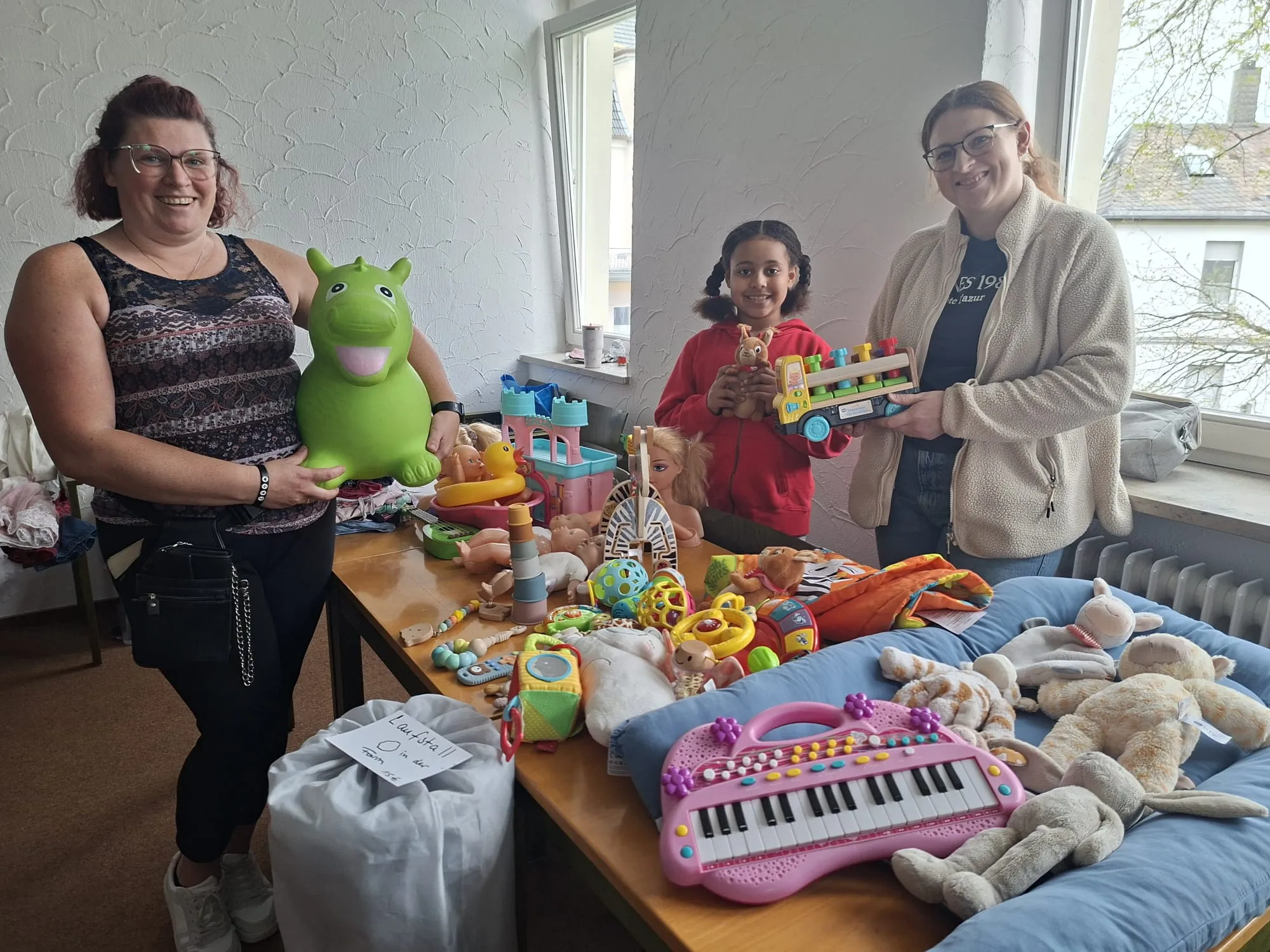 Drei Frauen stehen vor einer Auswahl an Kinderkleidung und Spielzeug. Links hält eine Frau ein Stofftier, in der Mitte ein Mädchen einen Holzbauklotz und rechts eine Frau weitere Spielsachen.