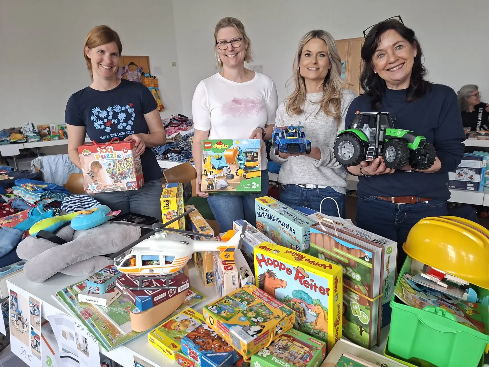 Vier Frauen stehen hinter einem Tisch voller Kinderkleidung, Spielzeug und Puzzles. Sie halten verschiedene Spielzeugautos in die Kamera.