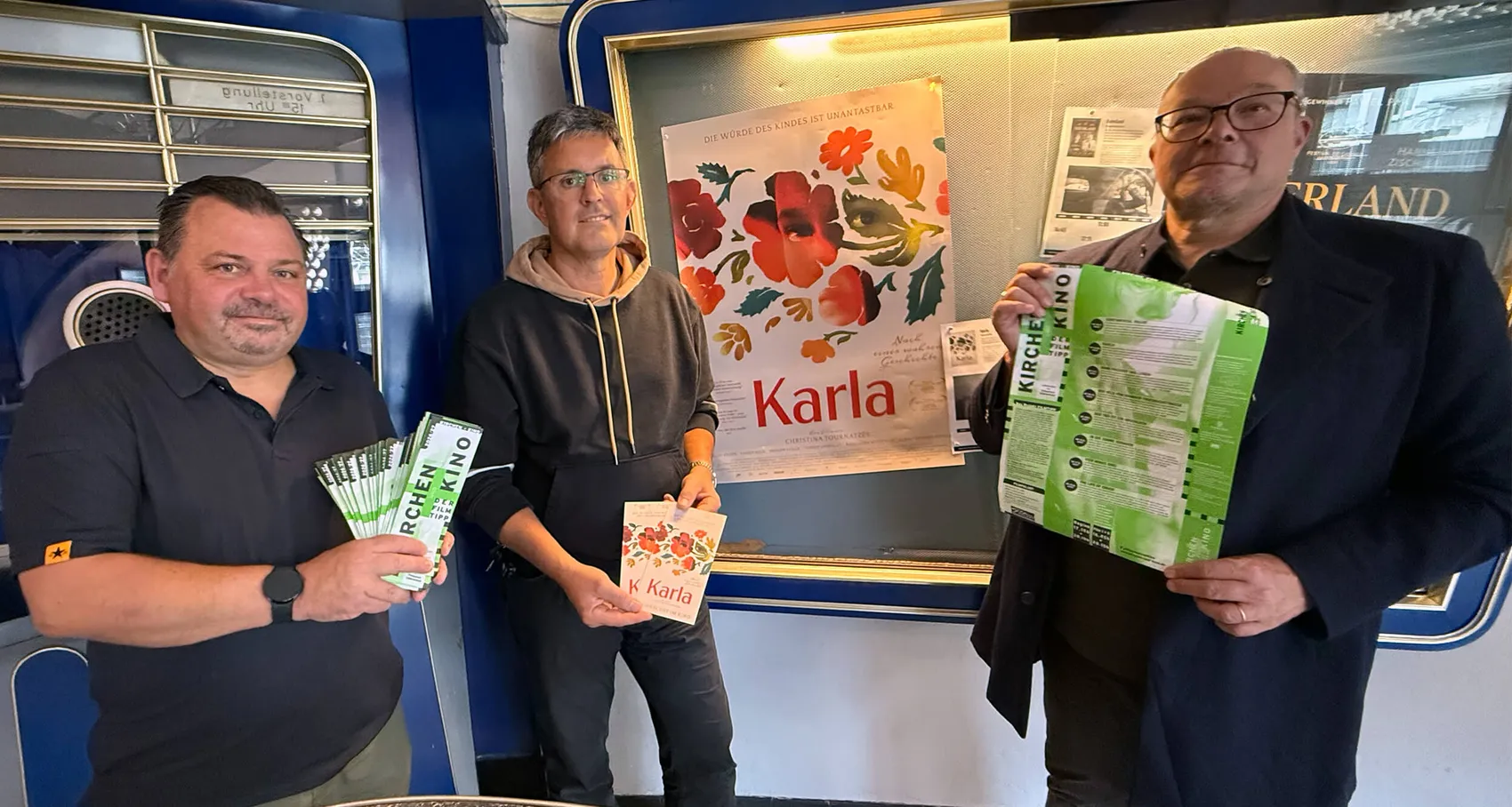 André Lubba, Günter Schmidt und Pfarrer Dirk Pollmann halten Flyer für die Filmreihe „Kirche und Kino“ vor einem Plakat des Films „Karla“.