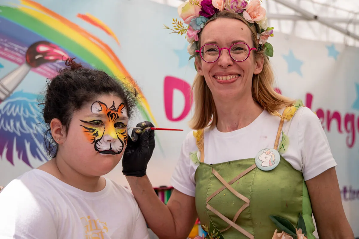 Eine Frau mit Blumenkranz und Tinkerbell-Kostüm bemalt das Gesicht eines Mädchens mit einem Tigerdesign.
