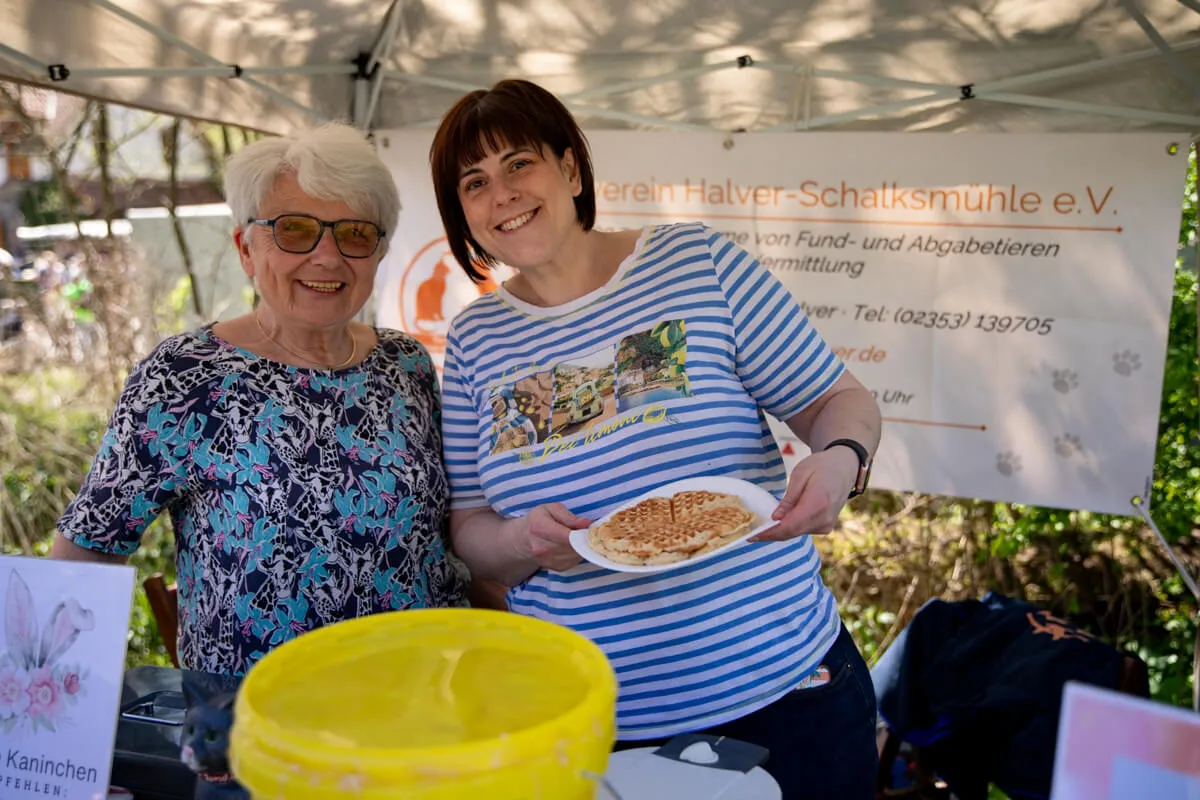 Zwei Frauen lächeln in die Kamera, eine hält einen Teller mit herzhaften Reibekuchen. Im Hintergrund ein Stand des Vereins Halver-Schalksmühle e.V.