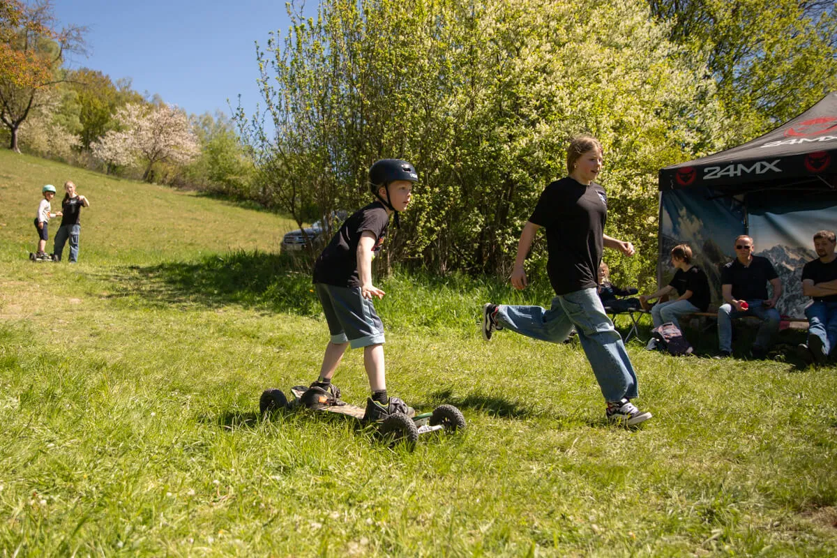 Zwei Jungen fahren auf einem grünen Feld mit Mountainboards, während im Hintergrund weitere Personen und ein Zelt zu sehen sind.