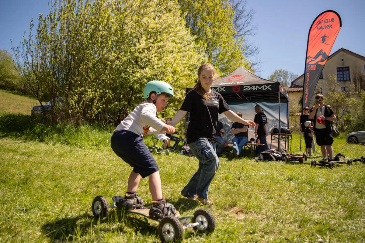 Ein Junge auf einem Mountainboard wird von einer jungen Frau begleitet, während sie auf einer Wiese fahren. Im Hintergrund steht ein Zelt und eine Flagge mit dem Schriftzug „Ski Club Halver“.