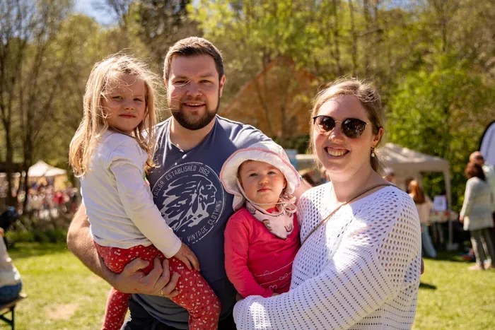 Familie mit zwei kleinen Kindern lächelt in die Kamera. Im Hintergrund Bäume und weitere Menschen auf einem Frühlingsfest.