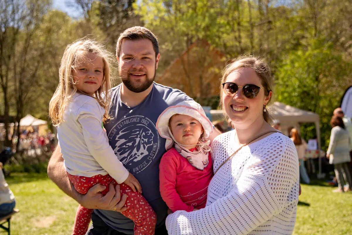 Familie mit zwei kleinen Kindern lächelt in die Kamera. Im Hintergrund Bäume und weitere Menschen auf einem Frühlingsfest.